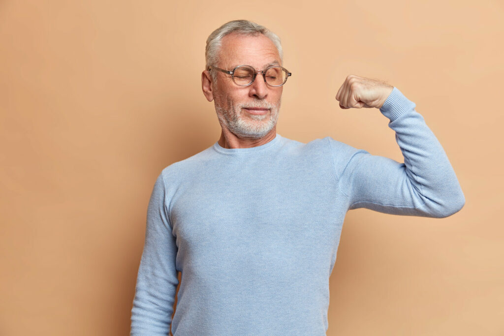 strong bearded grey haired man pensioner shows biceps stands with raised arm indoor wears jumper spectacles says look my strength demonstrates muscles isolated brown wall